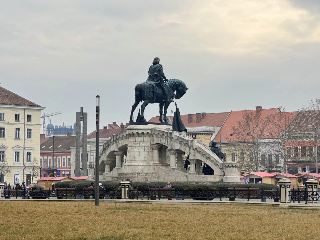 La imagen de la escultura por detrás con los edificios que rodean la plaza, de las más bonitas que sacar en Cluj-Napoca.